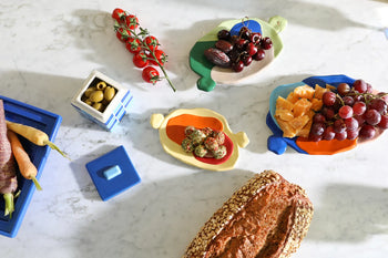 A &klevering Chunky Platter, Small displays seeded bread, fresh veggies, cherry tomatoes on the vine, cheeses, grapes, dates, and olives in a blue container—vivid snacks arranged with artistic charm by &klevering.