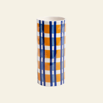 The &klevering Clash Check Vase by &klevering is a cylindrical ceramic vase with a glossy finish, showcasing bold hand-painted orange and blue plaid on white, styled against a plain light beige backdrop.