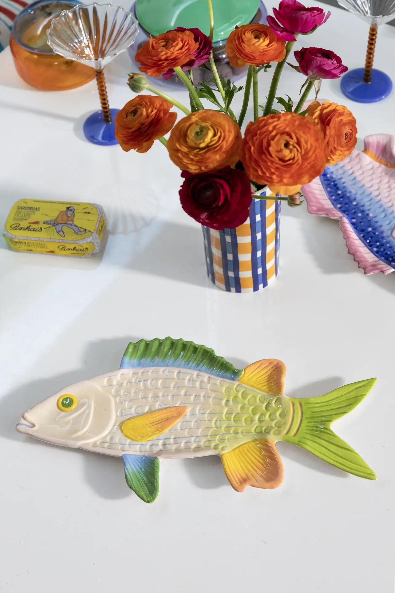 A ceramic fish plate with cheerful colours sits on a white table next to the &klevering Clash Check Vase filled with orange and red ranunculus. Hand-painted patterns, a tin of sardines, and colorful glassware add whimsy to the vibrant scene.