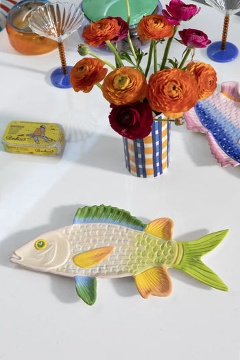 A ceramic fish plate with cheerful colours sits on a white table next to the &klevering Clash Check Vase filled with orange and red ranunculus. Hand-painted patterns, a tin of sardines, and colorful glassware add whimsy to the vibrant scene.