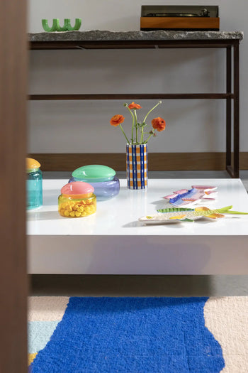 A white coffee table displays colorful glass containers, the &klevering Clash Check Vase in cheerful hues, and ceramic fish. Hand-painted patterns brighten the scene, with a blue and beige rug and a console table in the background.