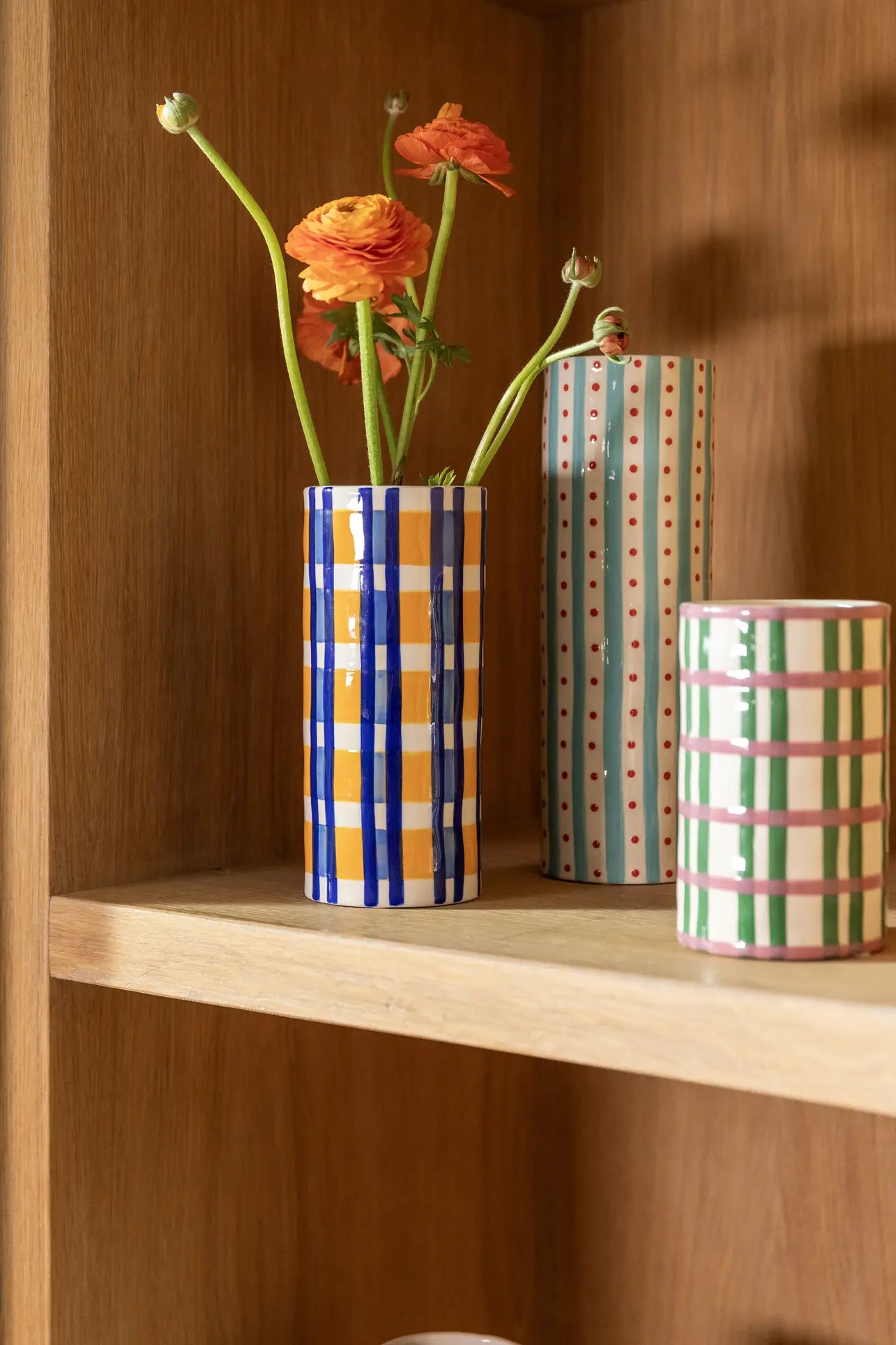The &klevering Clash Grid Vase by &klevering, featuring hand-painted colorful geometric patterns, holds orange flowers on a wooden shelf. Its vibrant stripes, checks, and dots add unique charm to the decor.