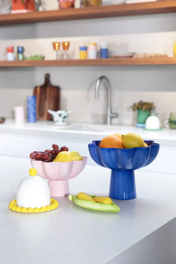 Colorful ceramic bowls with assorted fruit and a white butter dish rest on a modern kitchen counter, accompanied by the &klevering Corn Salt & Pepper Set; shelves, cups, and a sink can be seen in the background.