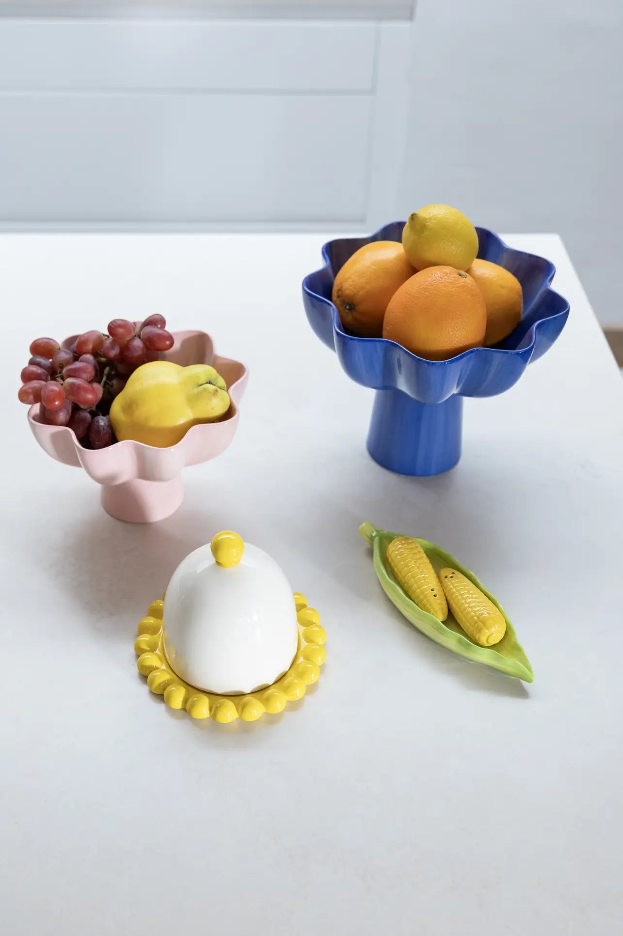 A kitchen dining table displays a pink bowl with grapes and quince, a blue bowl with oranges and lemon, a small covered dish with a yellow rim, and the &klevering Corn Salt & Pepper Set by &klevering in a ceramic pea pod.