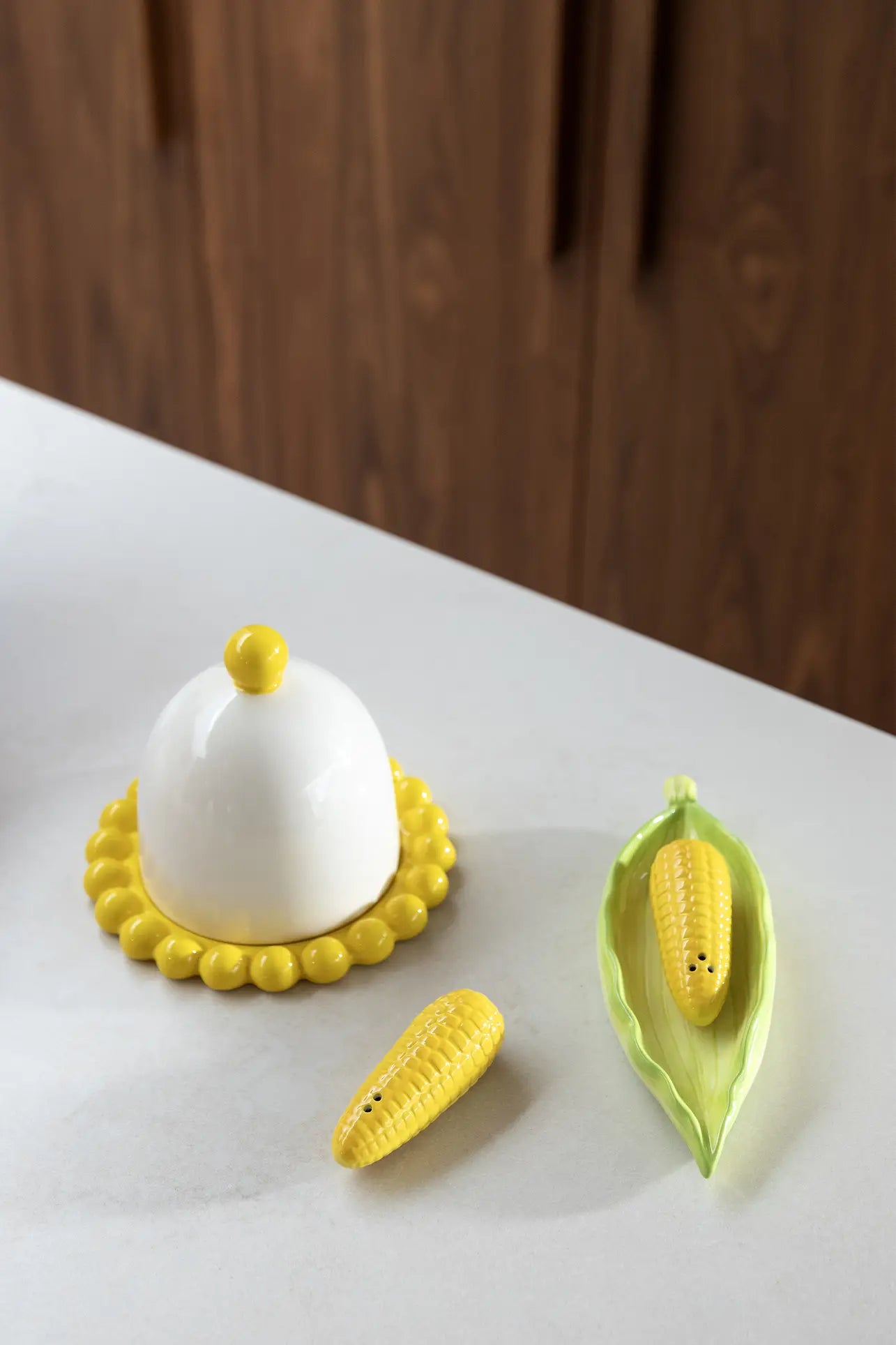 A white butter dish with a yellow beaded edge sits on the kitchen countertop beside the &klevering Corn Salt & Pepper Set, one shaker nestled in a green ceramic corn husk.