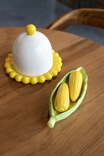 A ceramic butter dish with a yellow base and white dome lid sits on the table beside a corn husk-shaped tray holding the &klevering Corn Salt & Pepper Set from &klevering.