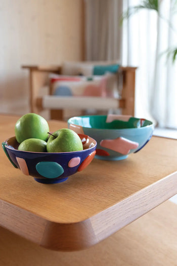 Two &klevering Crafty Bowls, Small—one holding green apples—rest on a wooden dining table in a bright, modern living room. Abstract shapes and patterned cushions accent the blurred background with a wooden sofa.
