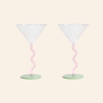 The &klevering Curved Coupe Glass Set of 2 features clear, scalloped rims, pink wavy stems, and green bases—charming colored glassware by &klevering, ideal for celebratory gatherings against a plain beige backdrop.