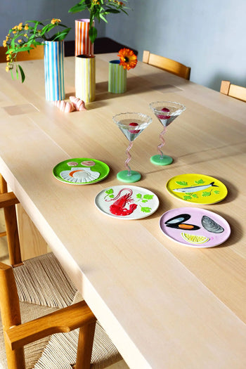 A light wooden dining table set for a celebration with four seafood-themed plates and two &klevering Curved Coupe Glasses, surrounded by wooden chairs and striped vases filled with flowers in the background.