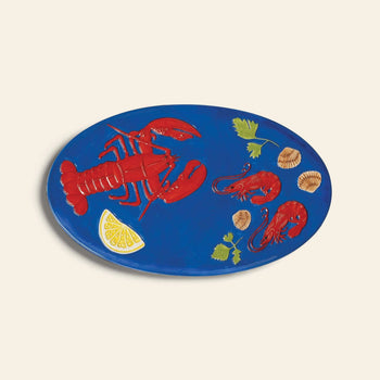 The &klevering De La Mer Lobster Platter is an oval blue platter with red lobster, shrimp, seashells, parsley leaves, and lemon wedge illustrations—ideal as a decorative piece for any coastal-themed table.