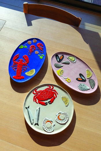 Three oval &klevering De La Mer ceramic plates rest on a wooden table, each adorned with vibrant seafood art—a Lobster Platter, one with mussels and oysters, and another with crab and fish—illuminated by soft sunlight.