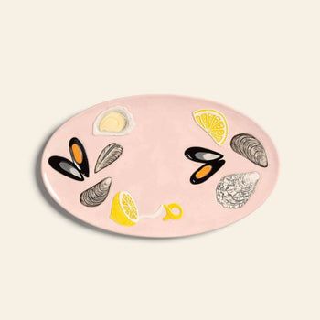 The &klevering De La Mer Oyster Platter is an oval pink platter featuring hand-painted illustrations of mussels, oysters, and lemon wedges, all by &klevering and set against a plain light background.