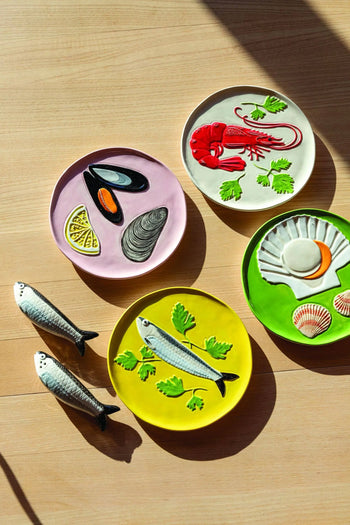 The &klevering De La Mer Plate Set of 4 features vibrant seafood designs—lobster, mussels, scallop shells, and sardines—adding oceanic charm to a light wood surface beside two ceramic sardine figurines in soft sunlight.