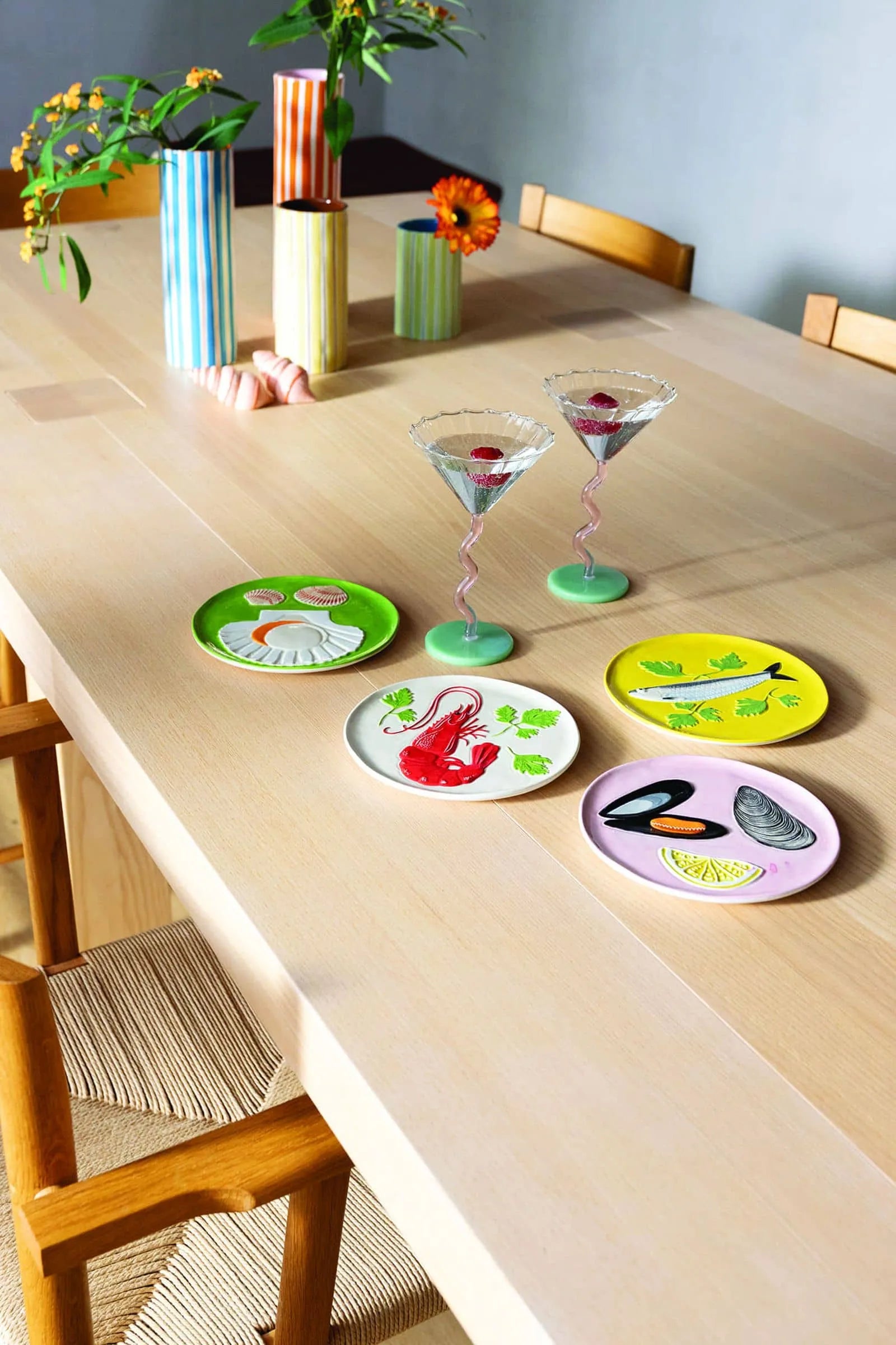 A light wooden dining table set with four &klevering De La Mer Plates, two martini glasses with pink drinks, and striped vases in the background brings oceanic charm to any meal.