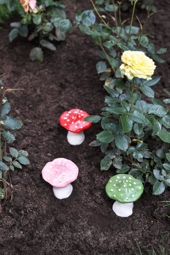 Three colorful &klevering Dots Mushroom Candles—red, pink, and green—stand in dark soil among rose bushes with a blooming yellow rose nearby, creating an enchanting garden ambiance.