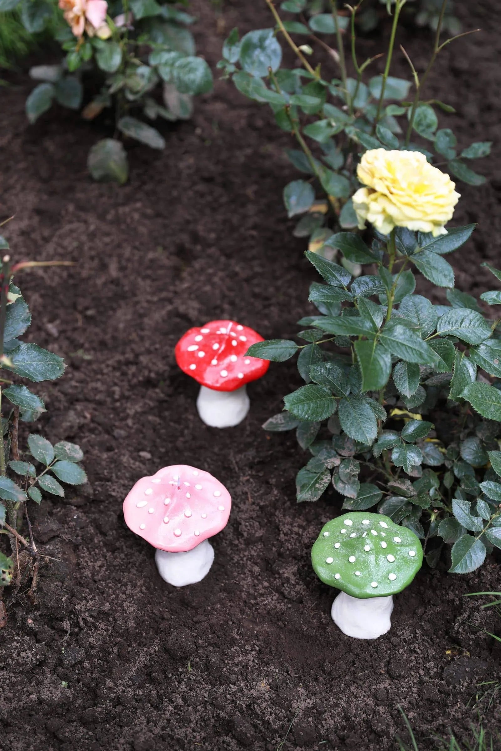 The &klevering Dots Mushroom Candle by &klevering adds charm to your garden with its colorful design, standing out among dark soil, green foliage, and blooming yellow roses.
