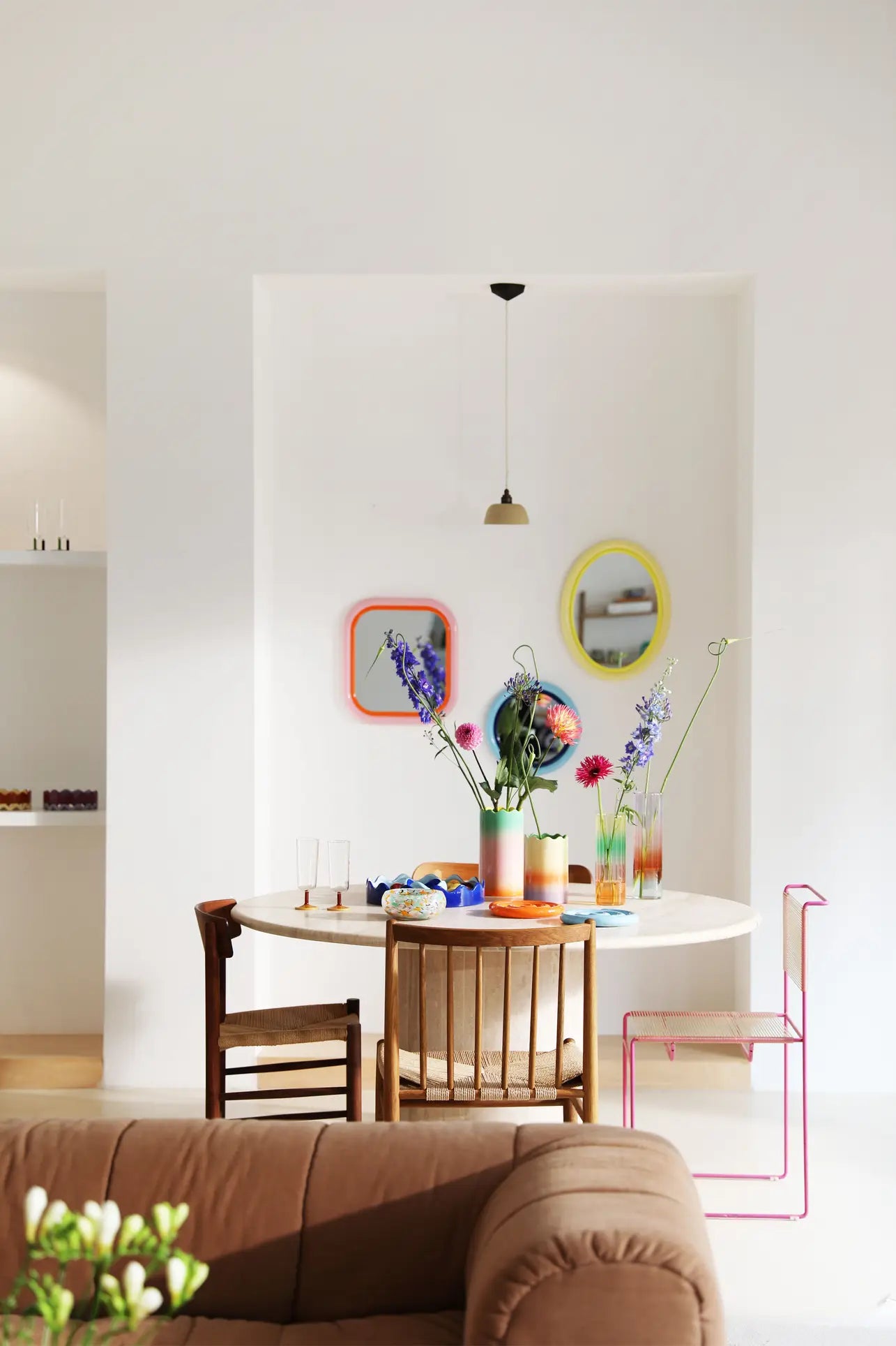 A cozy dining area features a round table styled with the &klevering Fade Vase, Large filled with colorful flowers, mismatched chairs, three pastel gradient oval mirrors on a white wall, and a glowing pendant light above.