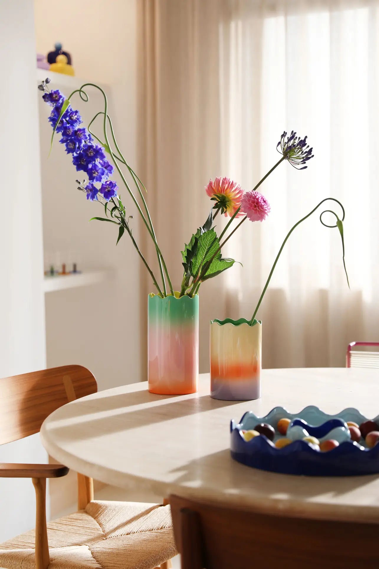Two &klevering Fade Vase, Large by &klevering, with wavy edges and pastel gradients, hold tall flowers on a round light wood table. Sunlight filters through sheer curtains; a blue bowl of marbles sits nearby with a wooden chair partly seen.