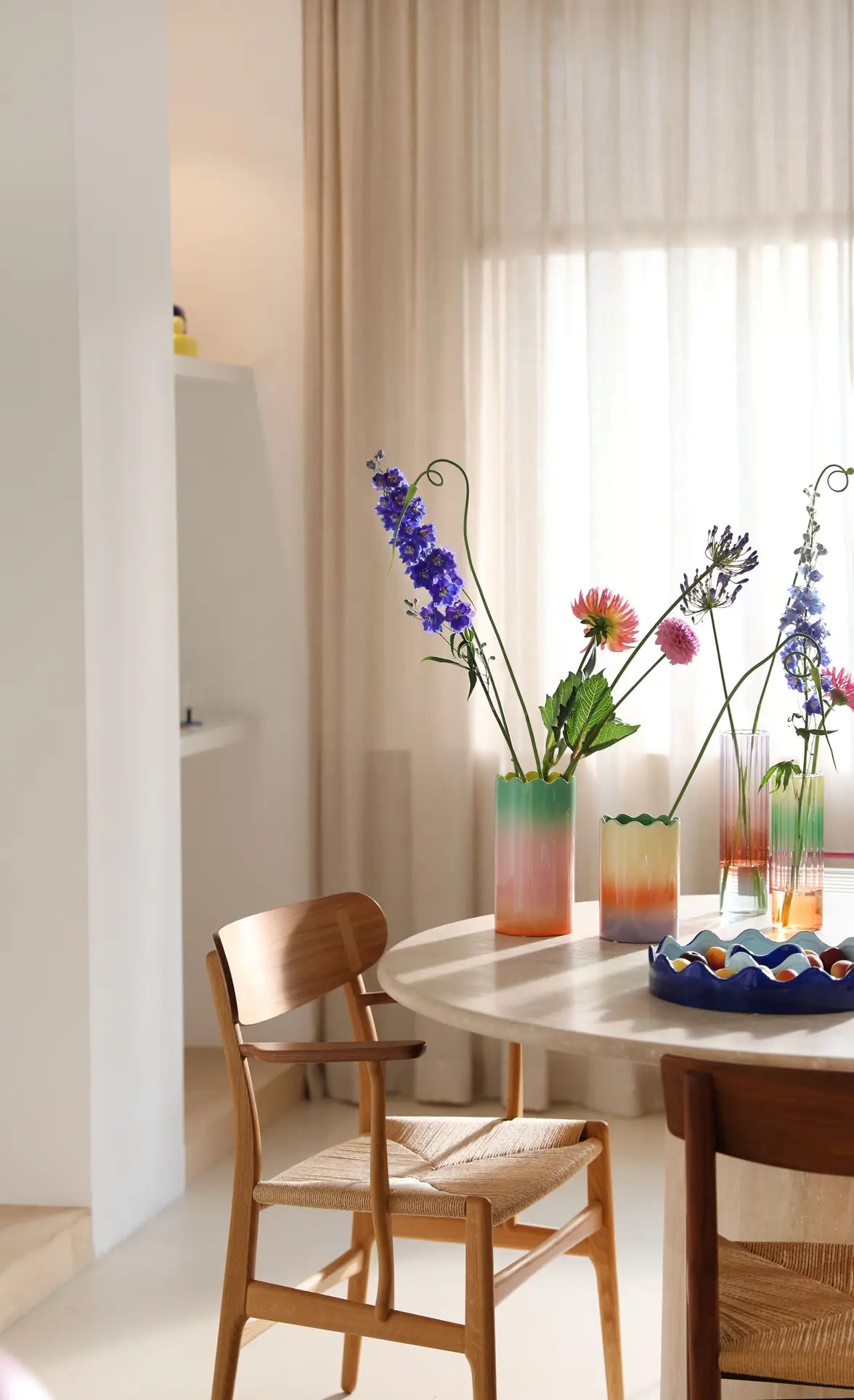 A bright dining area features wooden chairs, a round table with a pastel gradient tablescape, and an &klevering Fade Vase, Large by &klevering filled with fresh flowers under sunlight through sheer beige curtains for a warm, inviting feel.