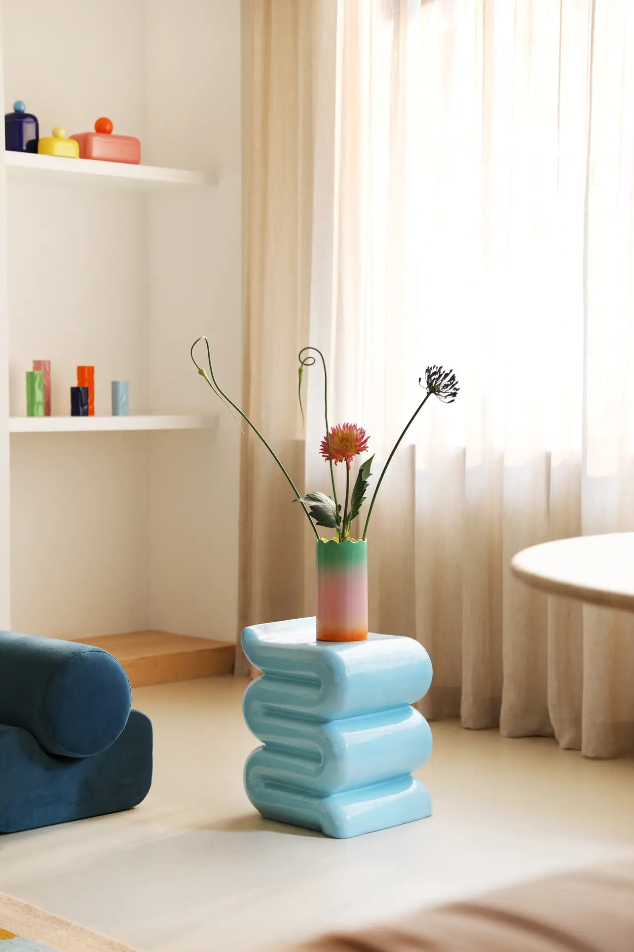 A pastel-toned living room features a blue wavy side table topped with the &klevering Fade Vase, Large by &klevering filled with colorful flowers. Soft sheer curtains and white shelves display decor in pastel gradients by the window.
