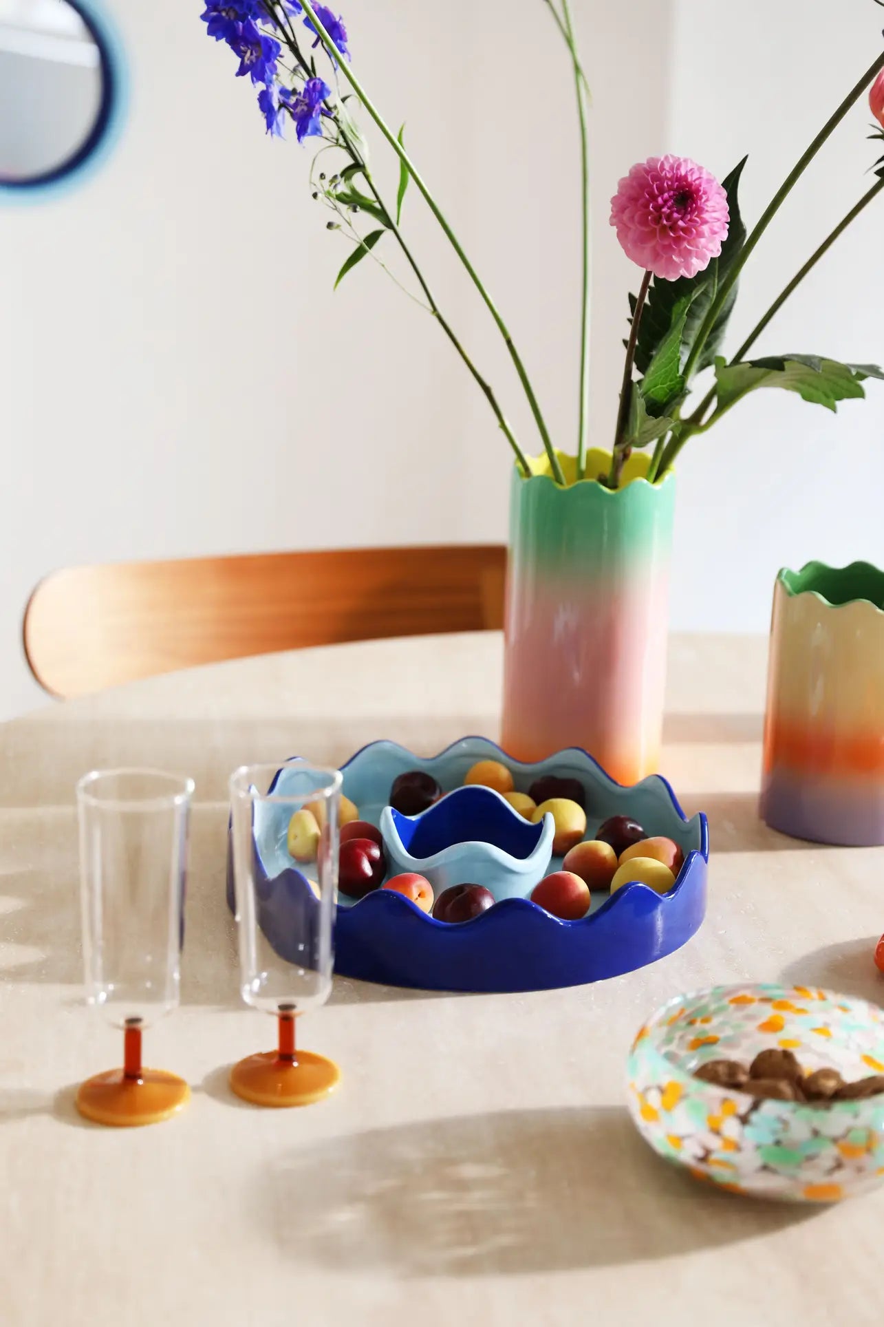 A round table with pastel gradient tones, vibrant glassware, a blue wavy bowl of fruit, two clear glasses with orange bases, a multicolored decorative plate, and an &klevering Fade Vase, Small by &klevering filled with flowers.