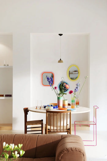 A modern dining area features a round table, mismatched chairs, and &klevering Fade Vases (Small) filled with flowers. Three oval pastel gradient mirrors brighten the white wall, with a pendant light above and a brown sofa in the foreground.