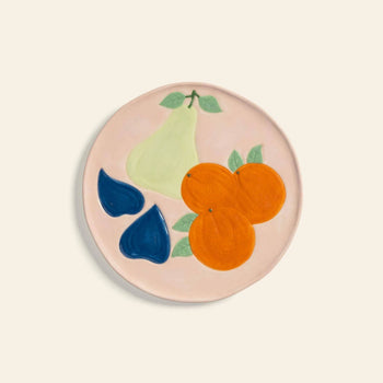 The &klevering Fruitful Plate, Set of 4 features vintage-inspired ceramic plates adorned with fruit motifs—a green pear, two oranges, and blue figs with leaves—on a pale pink and cream background.