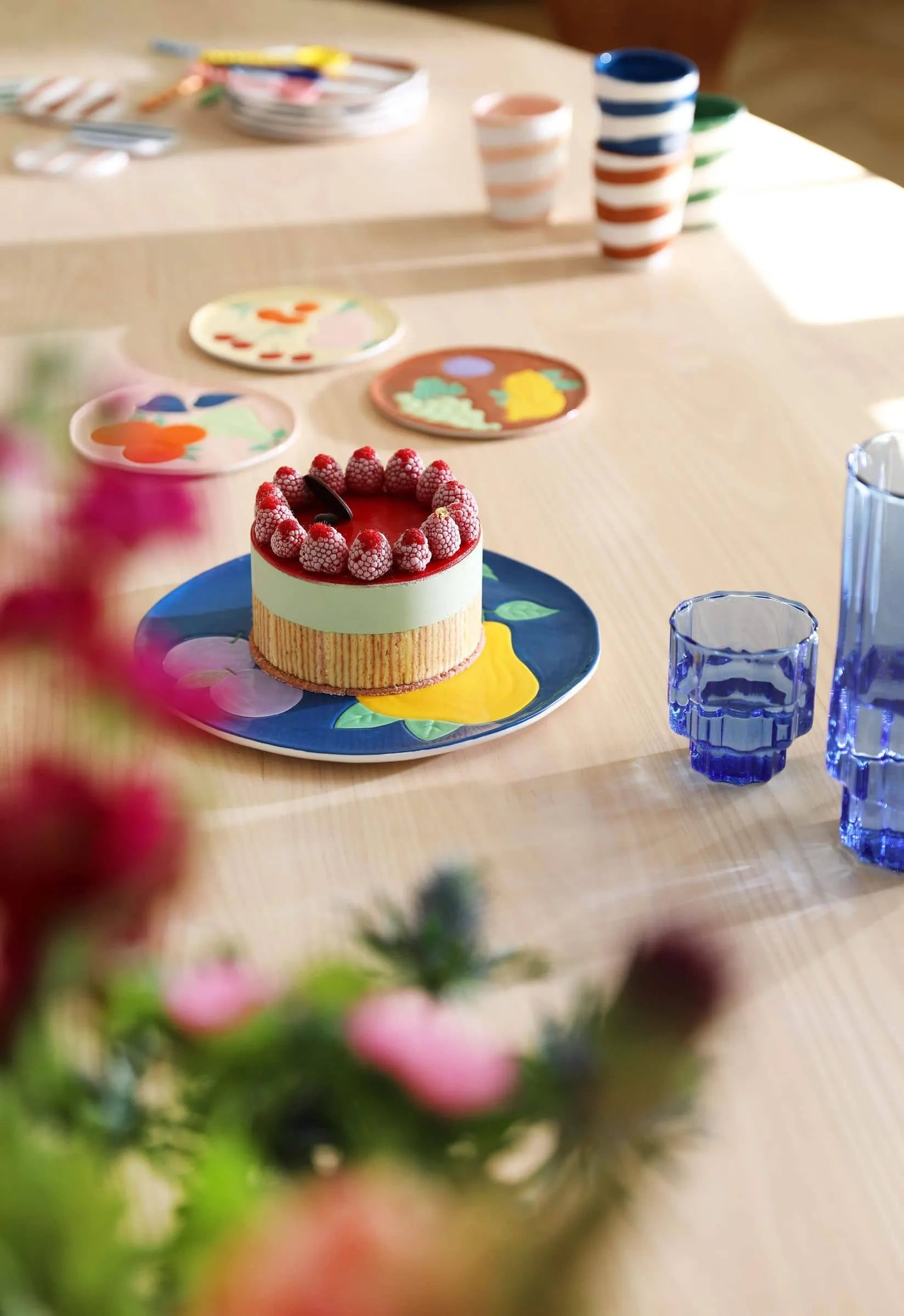 A small cake with strawberries sits on an &klevering Fruitful Plate from the set of 4 by &klevering, placed on a light wooden table with blue glasses, striped cups, and floral plates—evoking vintage charm amid blurred foreground flowers.