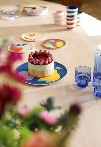 A small cake with strawberries sits on an &klevering Fruitful Plate from the set of 4 by &klevering, placed on a light wooden table with blue glasses, striped cups, and floral plates—evoking vintage charm amid blurred foreground flowers.