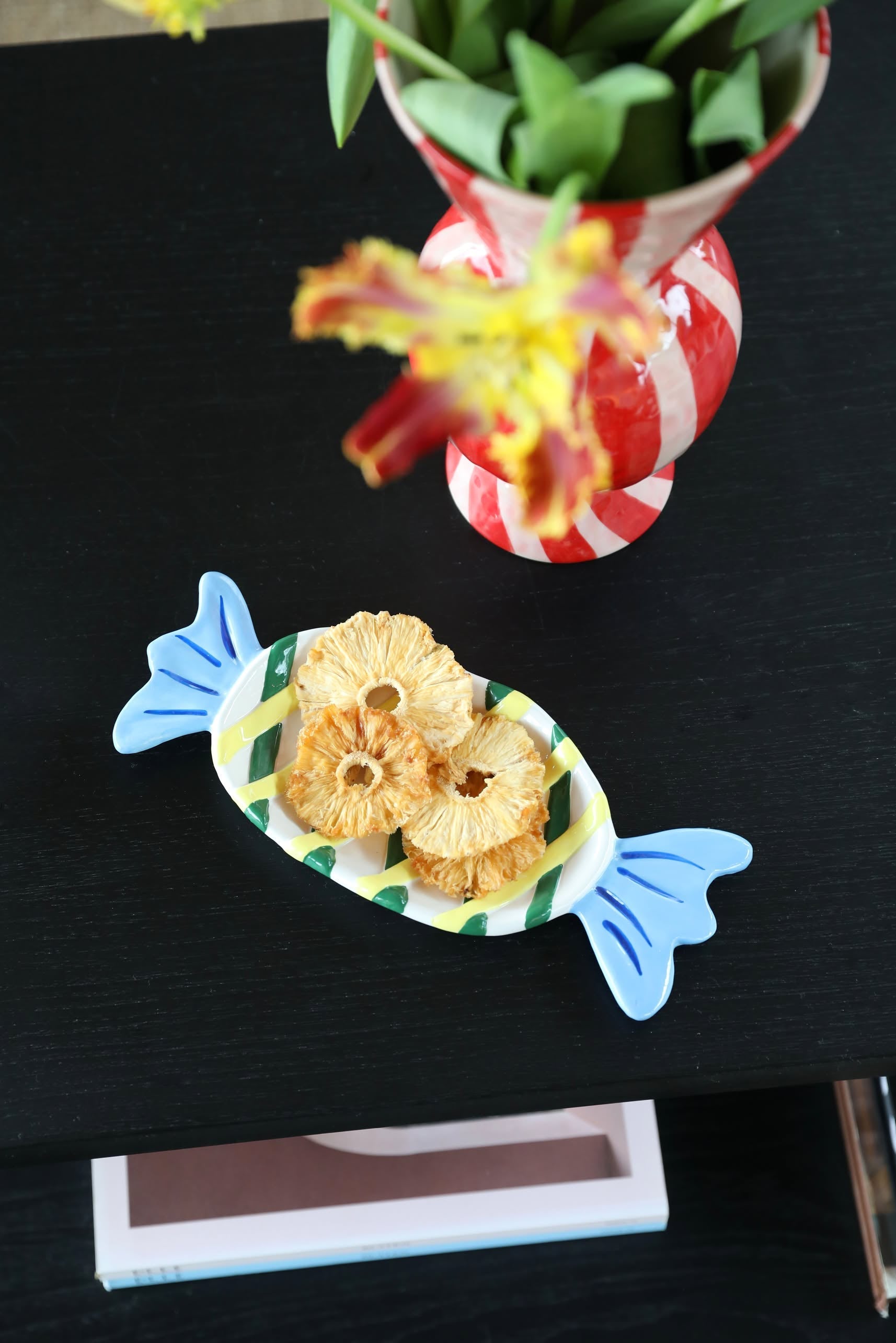 A medium &klevering Goody Plate with a whimsical design and blue ends holds dried pineapple rings on a dark table beside a red-and-white striped vase of yellow flowers.