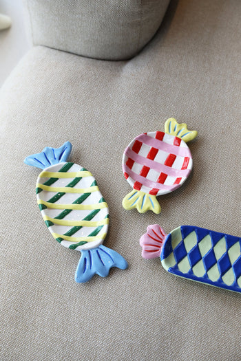 Three &klevering Goody Plates, Medium, by &klevering—ceramic plates shaped like wrapped candies with colorful checkered and striped designs—are arranged on a beige fabric surface.