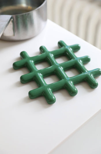 The &klevering Grid Trivet by &klevering, featuring a green glossy finish and geometric design, sits on a white surface next to a stainless steel pot, adding stylish heat protection.