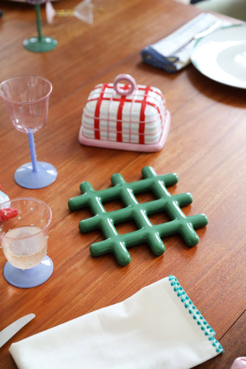 A wooden table set with pastel glassware, an &klevering Grid Trivet for heat protection with a geometric design, a checkered ceramic butter dish, a white napkin with blue stitching, and a partially visible plate.