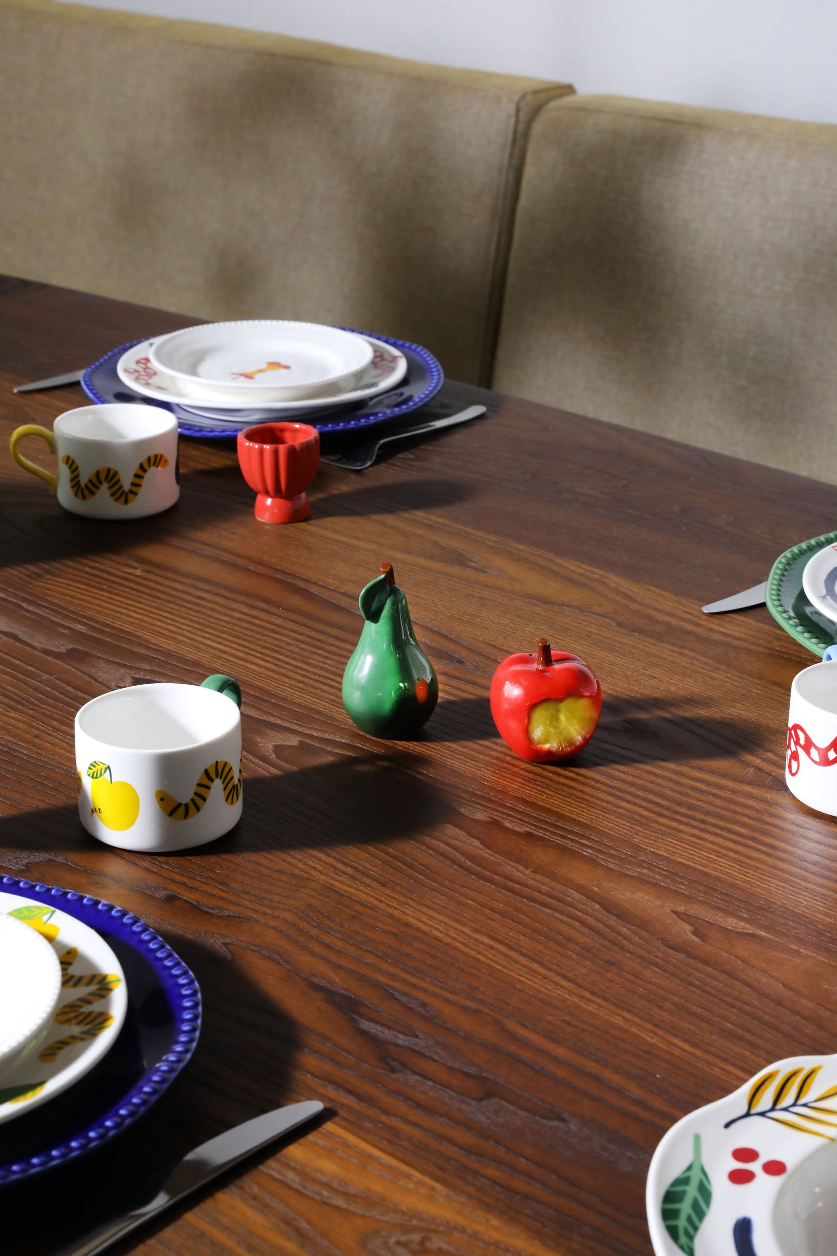 A wooden dining table set with colorful fruit-themed tableware is highlighted by the &klevering Haines Apple Pear Salt & Pepper Set and four stylish place settings, adding a fun and functional touch.