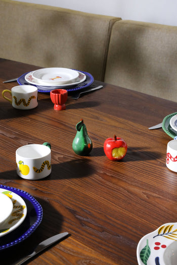 A wooden dining table set with colorful fruit-themed tableware is highlighted by the &klevering Haines Apple Pear Salt & Pepper Set and four stylish place settings, adding a fun and functional touch.