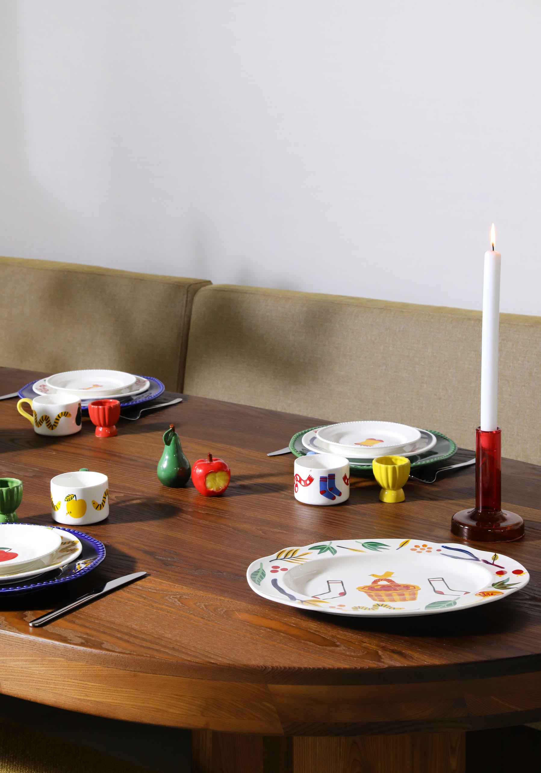 A wooden dining table set with colorful, patterned tableware, cups, and the &klevering Haines Apple Pear Salt & Pepper Set glows by a lit white candle in a red holder, with cushioned seating enhancing the playful atmosphere.
