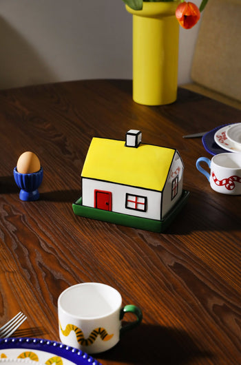 The &klevering Haines Butter Dish, with its house-inspired yellow roof design, rests on a wooden table surrounded by plates, a blue egg holder, and tulips—adding charm to any English breakfast setting.