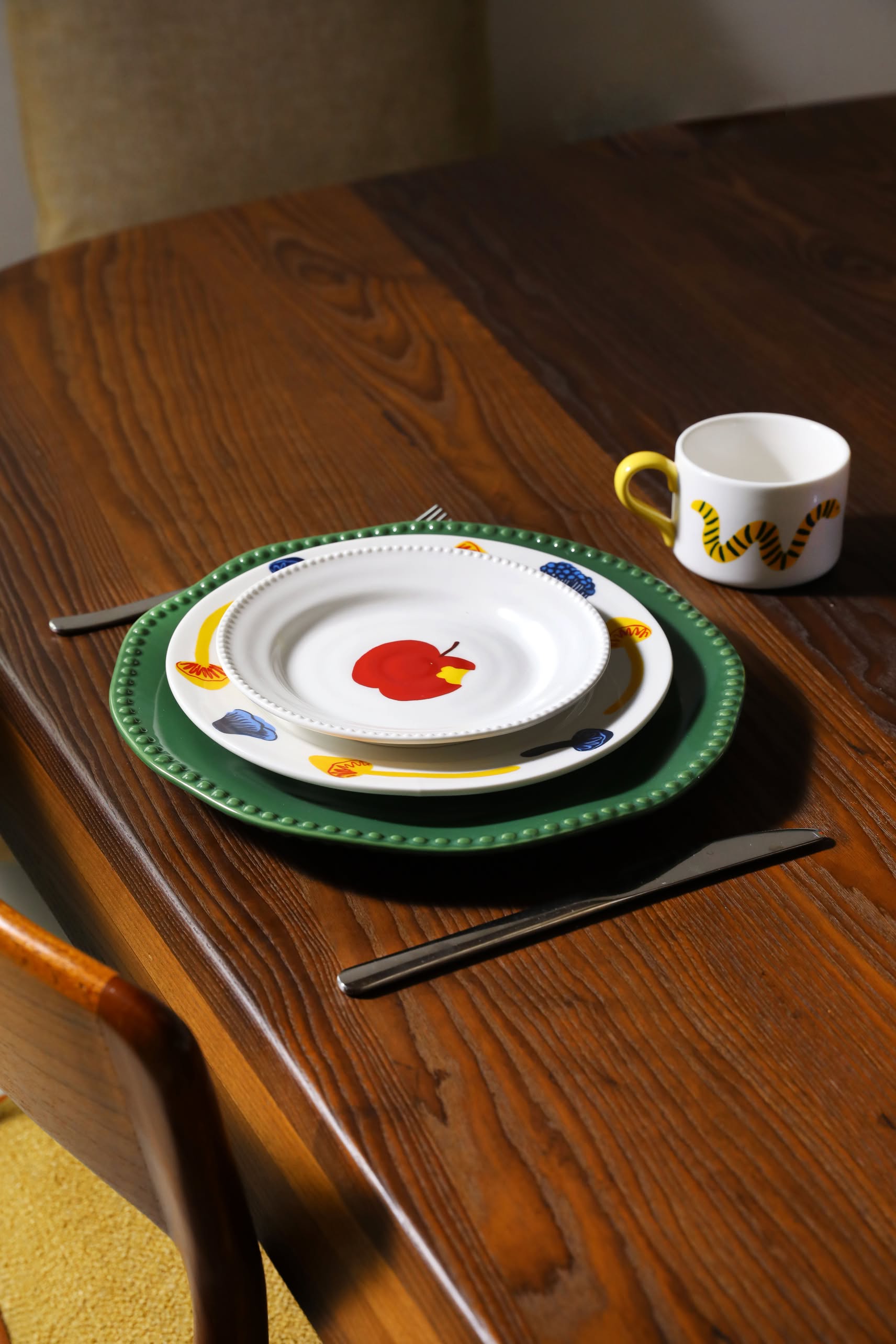 A wooden table set with the whimsical &klevering Haines Plate Small, Set of 4: green and illustrated plates, paired with cutlery and a mug featuring a yellow tiger stripe handle for a playful dining experience.