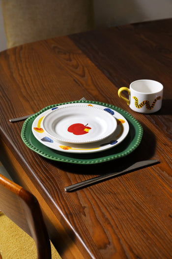A wooden table set with the whimsical &klevering Haines Plate Small, Set of 4: green and illustrated plates, paired with cutlery and a mug featuring a yellow tiger stripe handle for a playful dining experience.