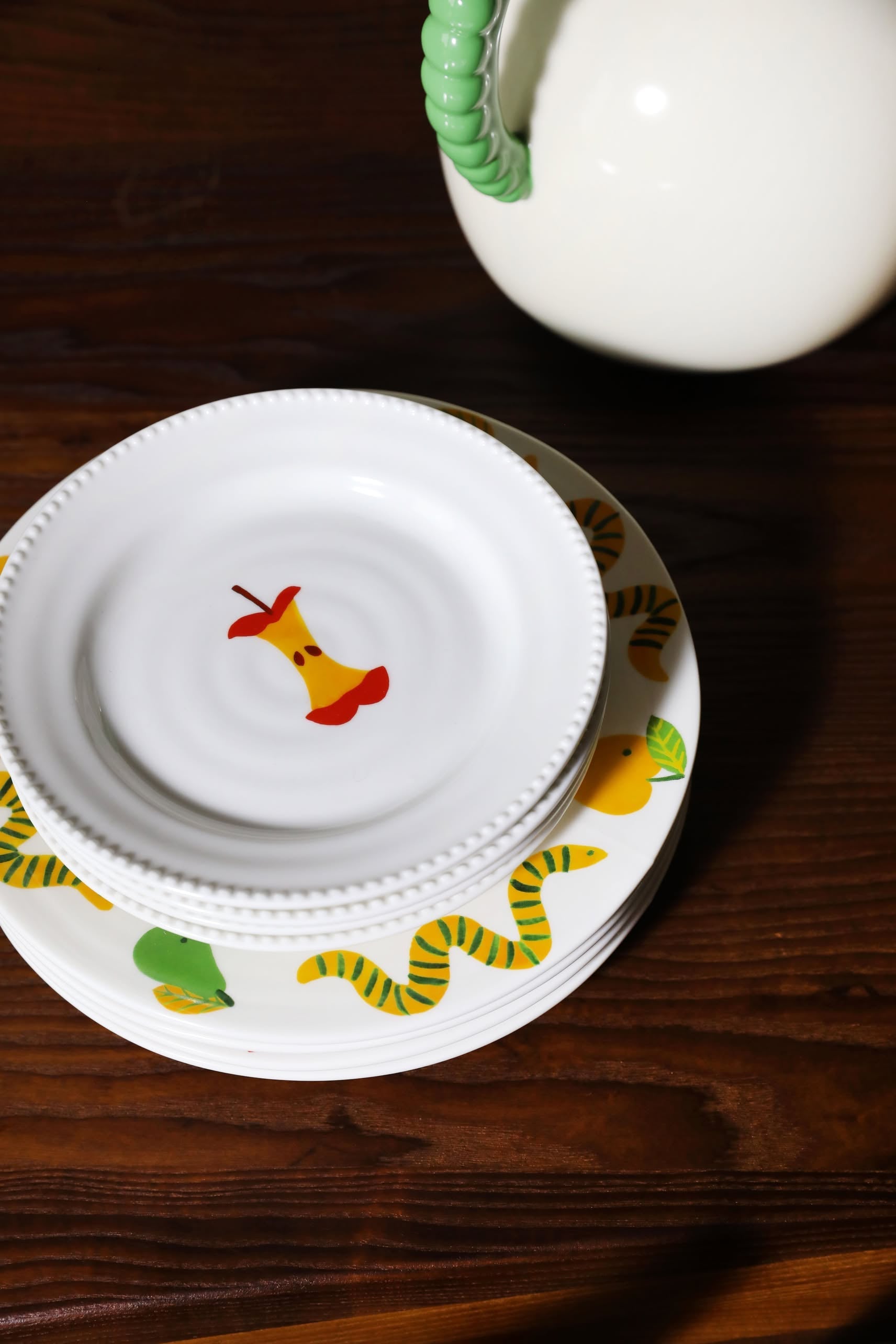 Two stacked white plates from the &klevering Haines Plate Small, Set of 4; the top plate displays a yellow apple core with red stem, and the bottom features a playful green and yellow striped snake—perfect for elevating your table.