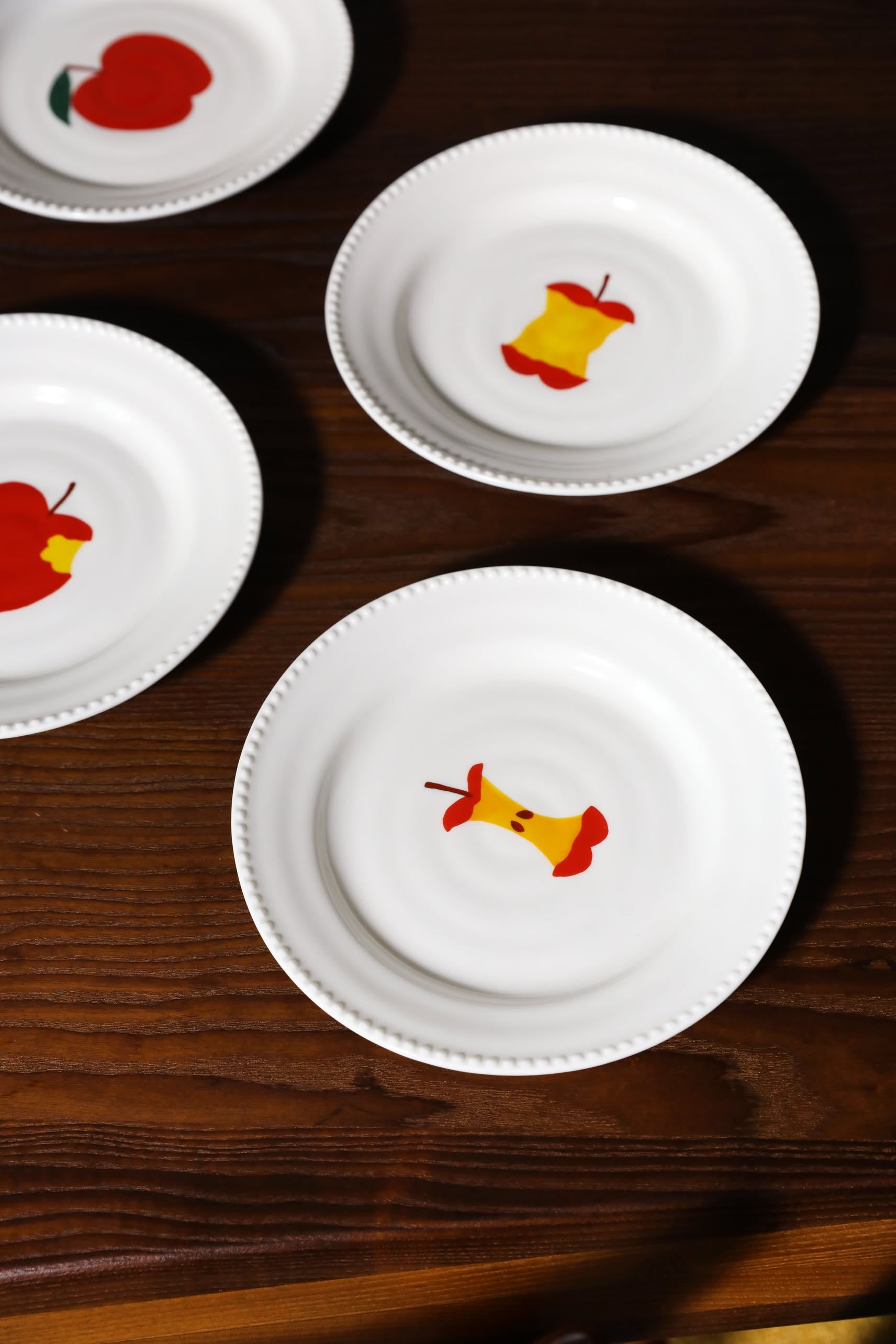 Three white &klevering Haines Plate Small plates, each with scalloped edges and a hand-painted apple or apple core, from the set of 4 by &klevering, are arranged on a dark wooden surface.