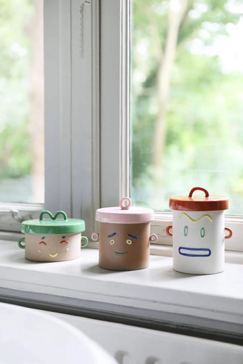Three &klevering Happy Jar containers with playful cartoon faces and handles by &klevering rest on a windowsill. Each unique in color and expression, they offer cheerful, versatile storage that brightens any space against the outdoors.