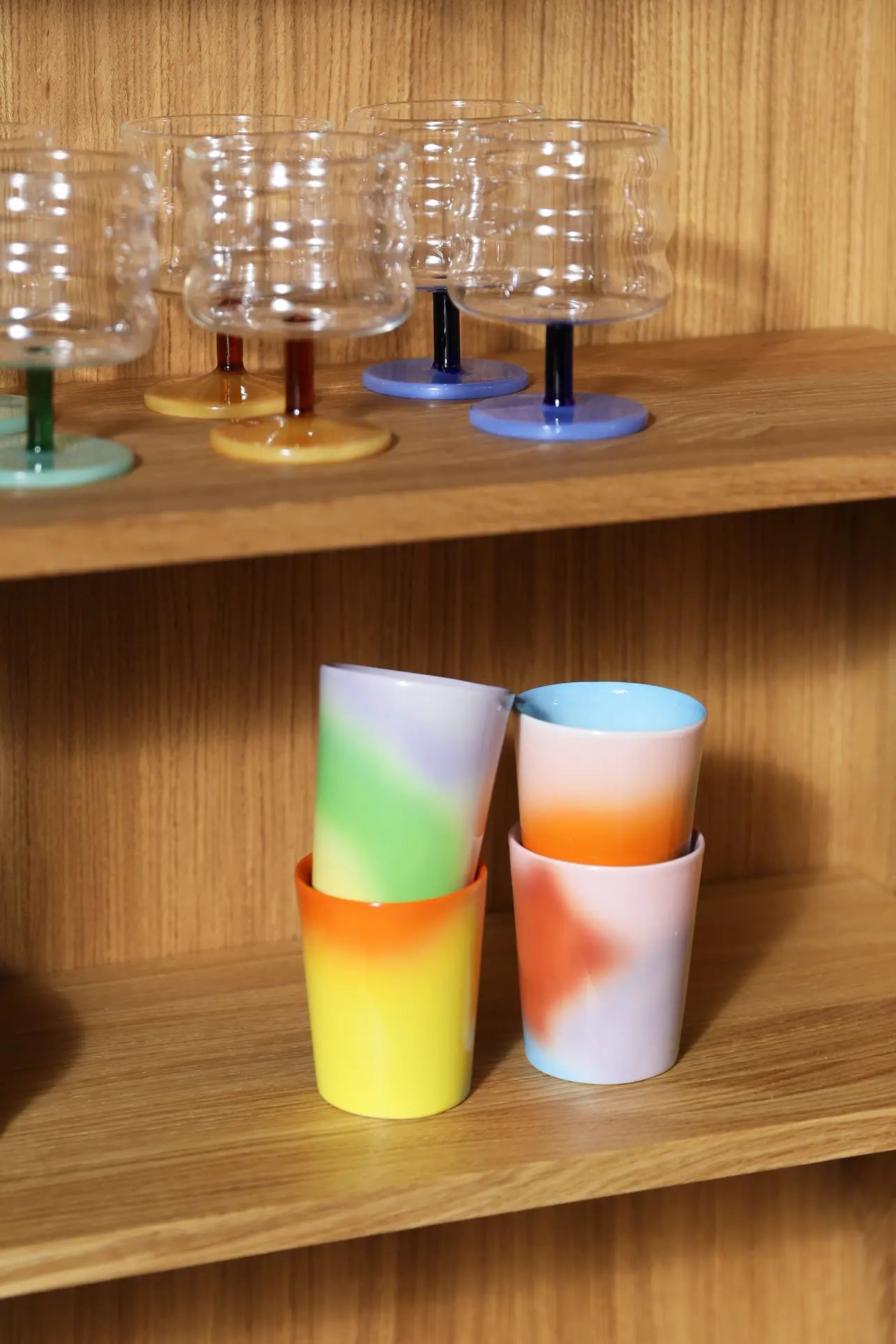 The &klevering Hue Mug Small, Set of 4 by &klevering features colorful mugs with wavy pastel hues displayed on a wooden shelf, with clear ribbed goblets featuring vibrant stems arranged above.