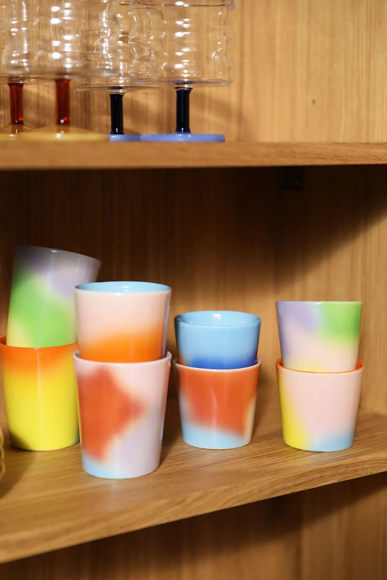 &klevering Hue Mug Small, Set of 4, featuring vibrant tie-dye patterns, are stacked on a wooden shelf. Above them, clear glass goblets with blue stems bring an elegant accent to the &klevering display.