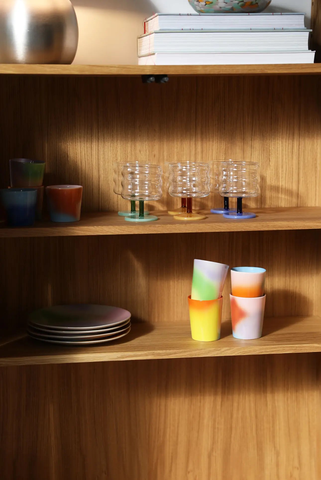 A wooden shelf displays a set of 4 &klevering Hue Mug Small mugs, clear ribbed glasses with drinks, stacked plates, and a few books on the upper shelf, all highlighted by warm lighting.