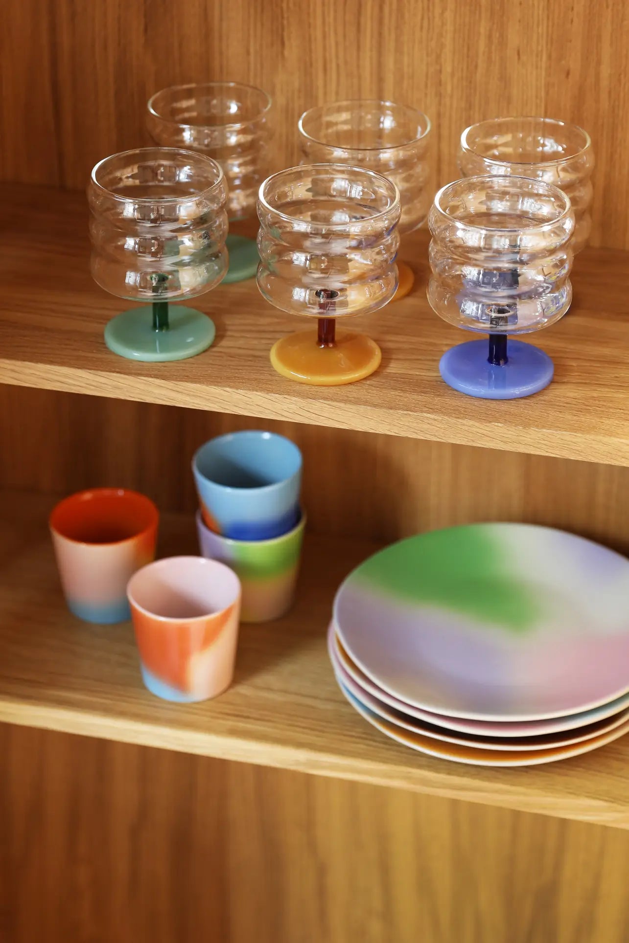 Colorful glassware with pastel bases sits on the top wooden shelf, while &klevering Hue Mug Small, Set of 4 and gradient plates in pastel hues are neatly arranged below against a wooden cabinet backdrop.