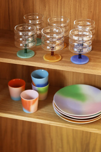 Colorful glassware with pastel bases sits on the top wooden shelf, while &klevering Hue Mug Small, Set of 4 and gradient plates in pastel hues are neatly arranged below against a wooden cabinet backdrop.