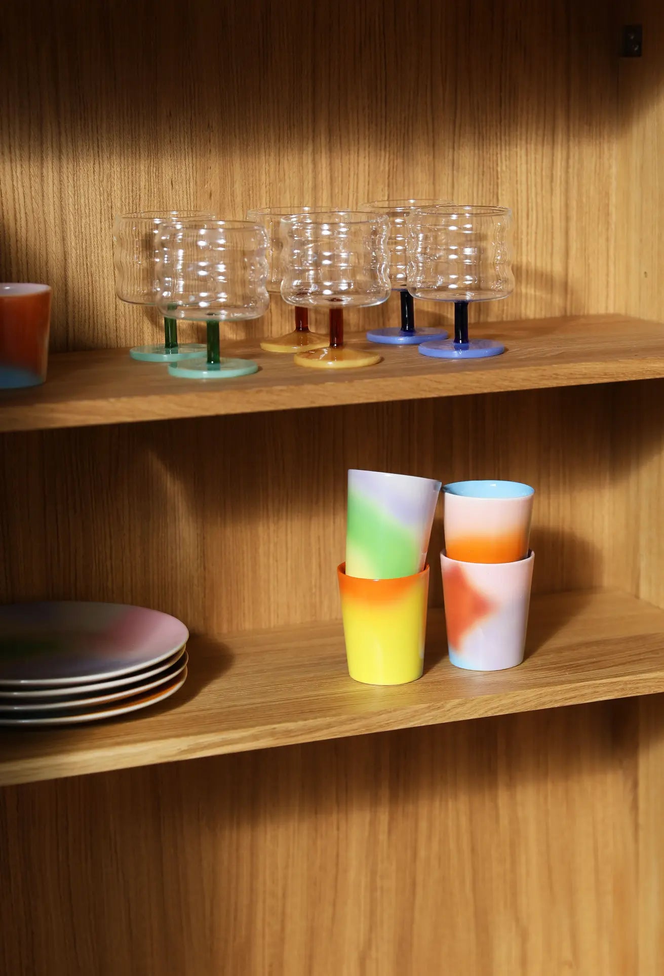 &klevering Hue Mug Small, Set of 4, are stacked on a wooden shelf, while a row of clear, textured glasses with colored stems sits on the shelf above.