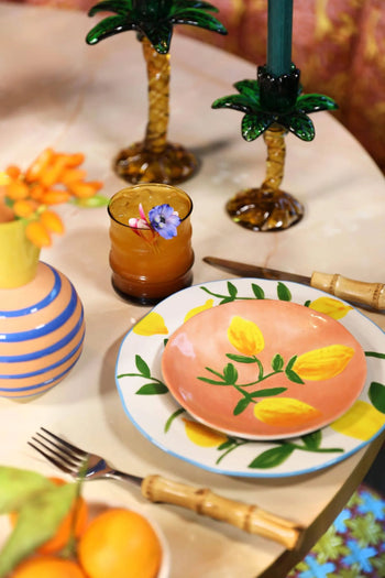 A colorful summer table features the &klevering Lemon Plate, Set of 4—playful plates with yellow lemons—plus a striped vase of orange flowers, an amber glass with a floral garnish, and palm tree candlesticks for vibrant style.