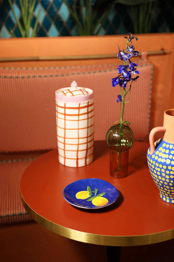 A round red summer table showcases &klevering Lemon Plates (set of 4), a patterned ceramic jar, a brown vase with purple flowers, and a jug with blue and yellow polka dots, set against a pink striped bench.